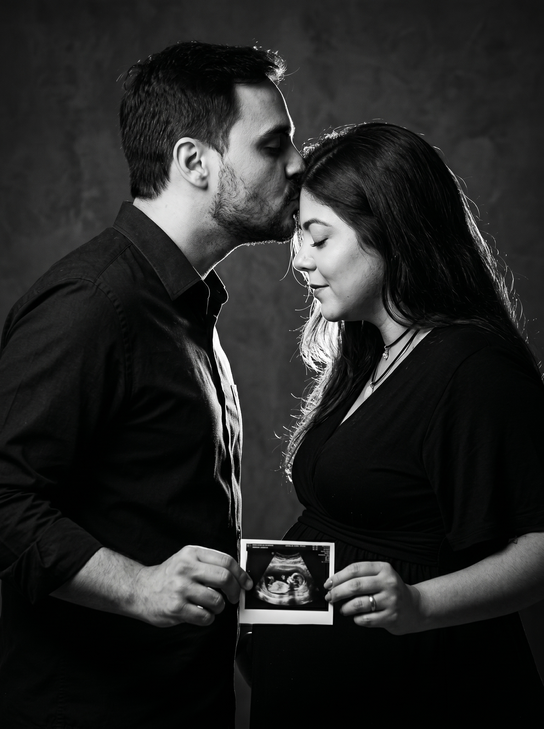 Casal de perfil em estúdio escuro, o homem beija a testa da mulher enquanto ambos olham para o ultrassom iluminado lateralmente.