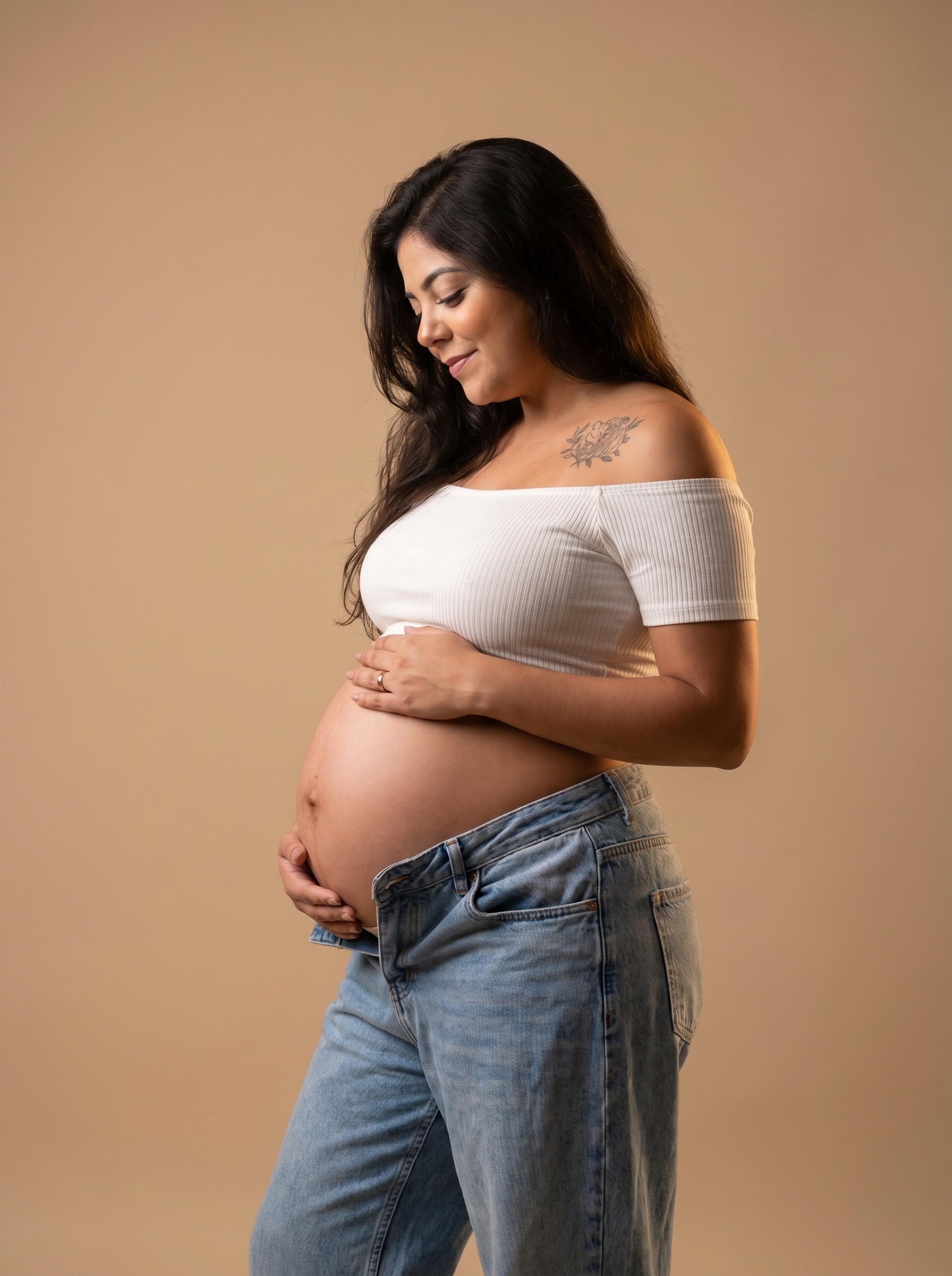 Mulher grávida de perfil em fundo bege, vestindo top branco e jeans, abraçando suavemente a barriga com ambas as mãos e olhando para baixo em um momento sereno.