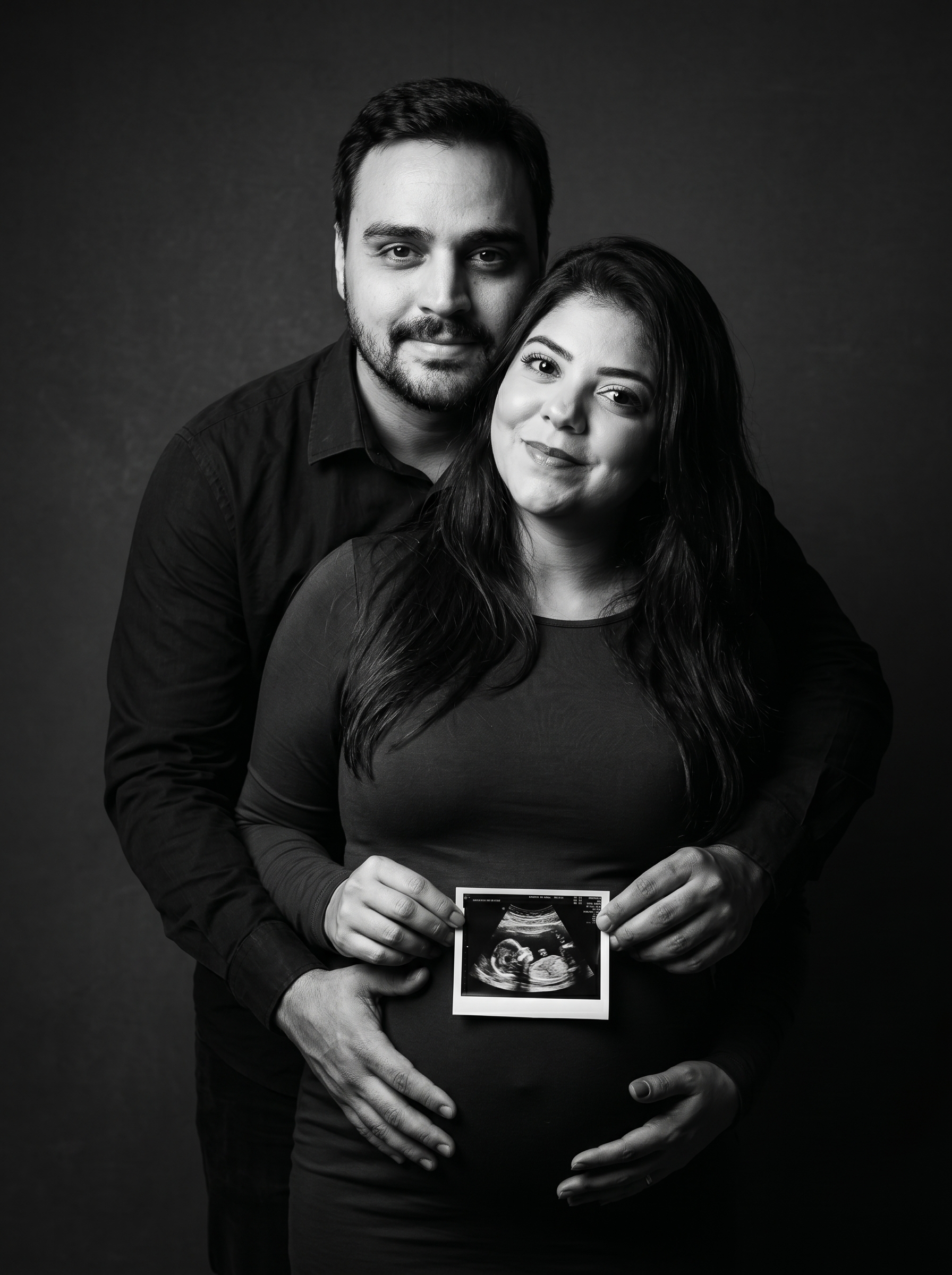Um homem abraça uma mulher grávida por trás em um estúdio escuro; ambos sorriem para a câmera enquanto seguram o ultrassom sobre a barriga da mulher.