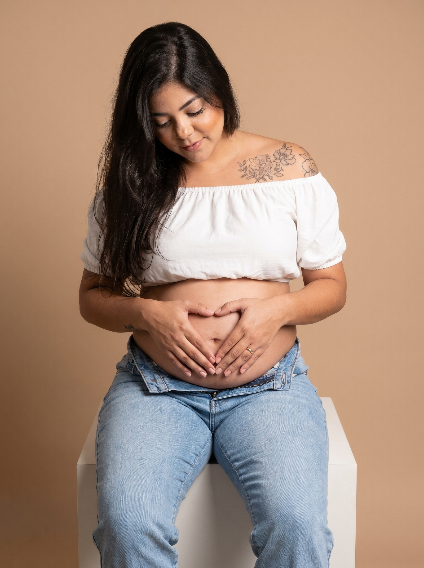 Close-up de uma mulher grávida sentada, com o enquadramento focado do peito aos joelhos, enquanto suas mãos formam um coração sobre a barriga exposta.