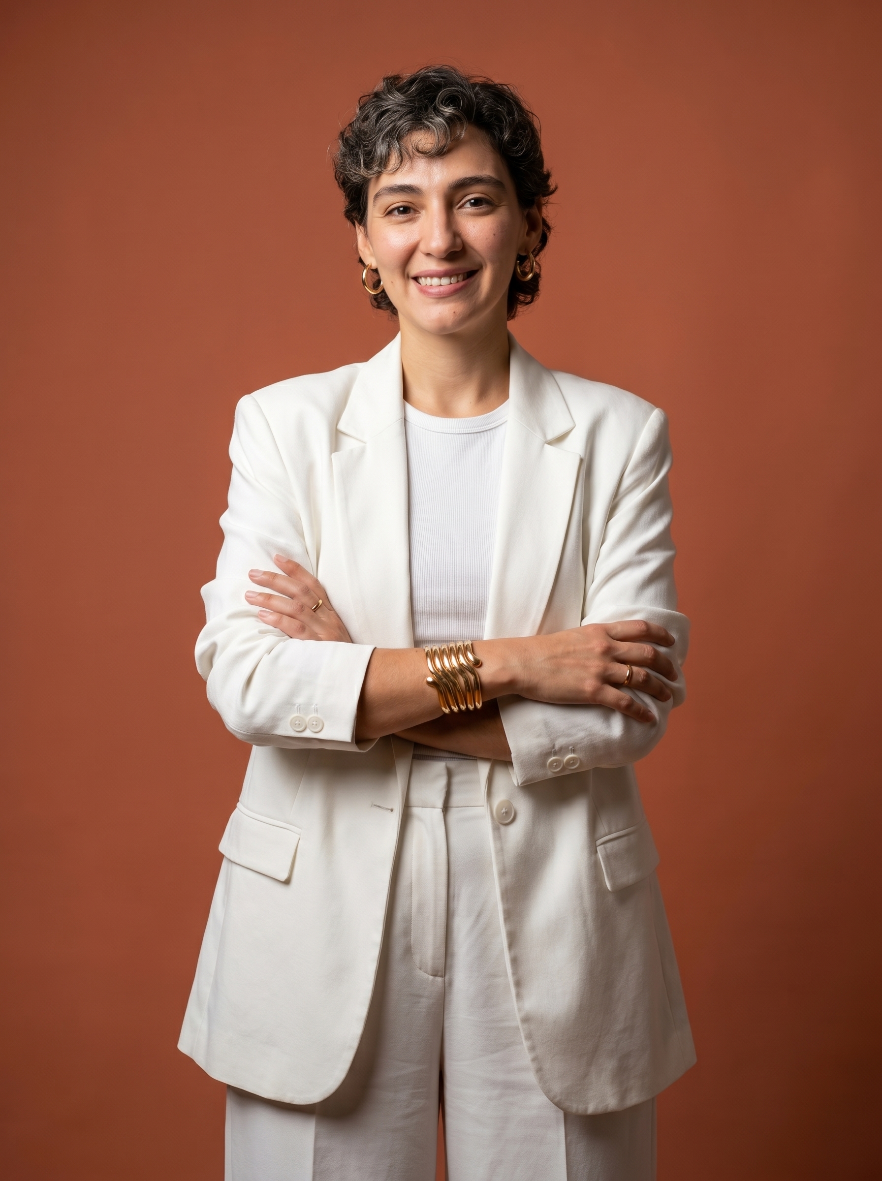 Mulher de blazer branco de pé, braços cruzados na altura do peito, postura aberta e confiante, leve sorriso, fundo terracota de estúdio.